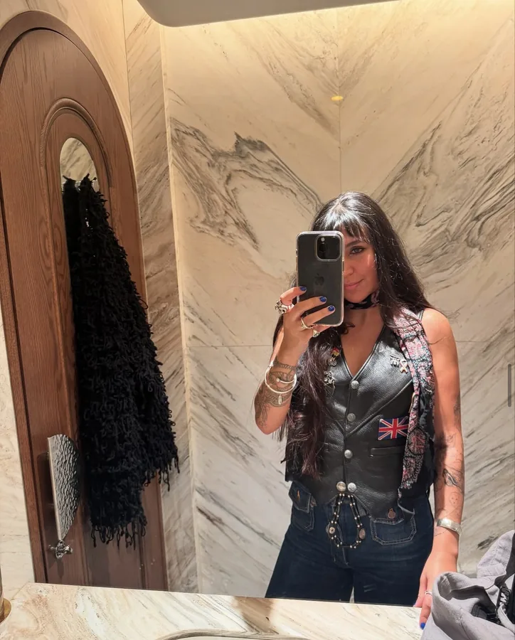Lily Rivers in a leather vest, photographing herself in a marble mirror
